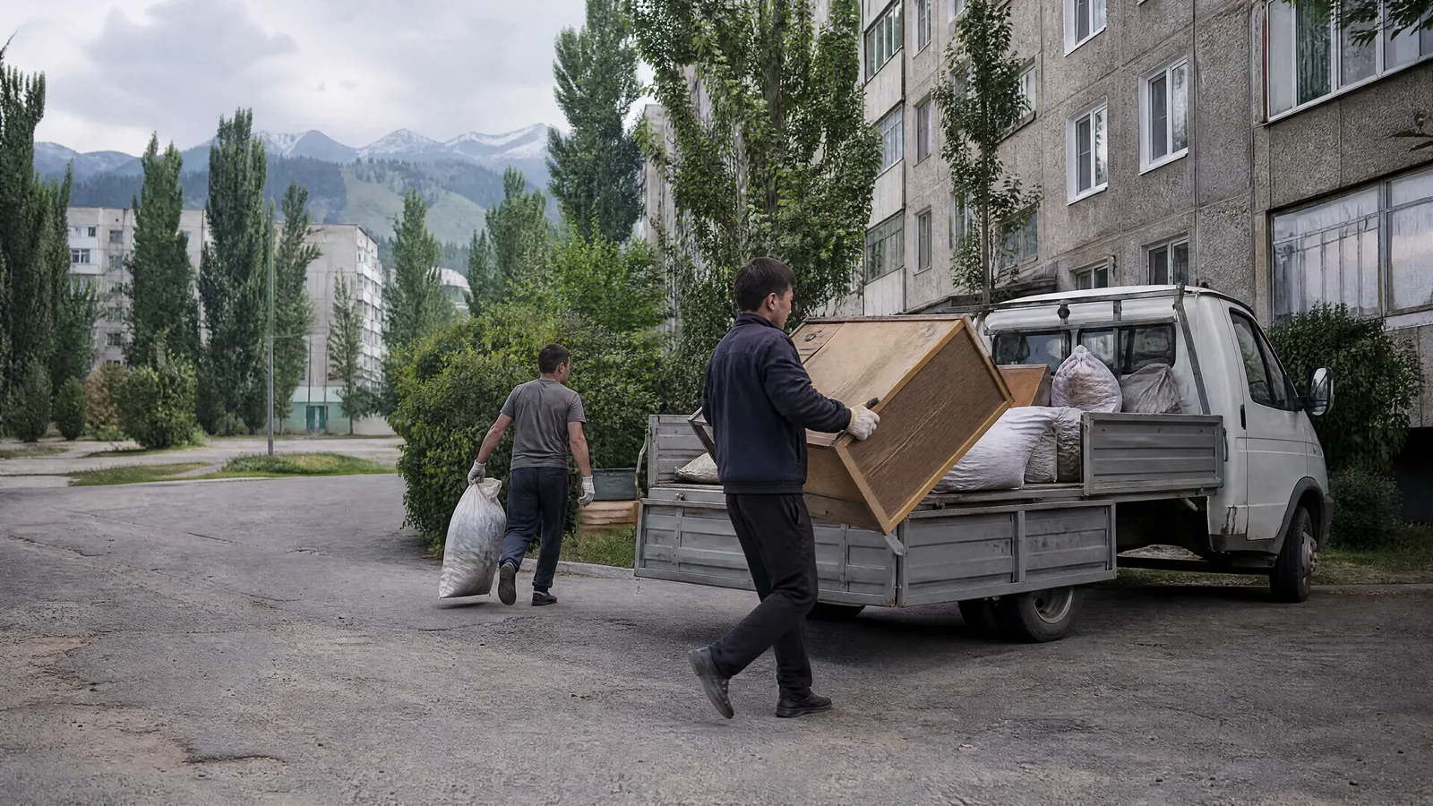 Двое грузчиков в перчатках несут старую мебель и мешки со строительным мусором к газели во дворе алматинской многоэтажки